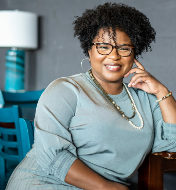 Erika Seth Davies, founder of the Racial Equity Asset Lab, sitting in a chair wearing a gray sweater and short natural hair smiling at the camera with her left hand slightly under her chin